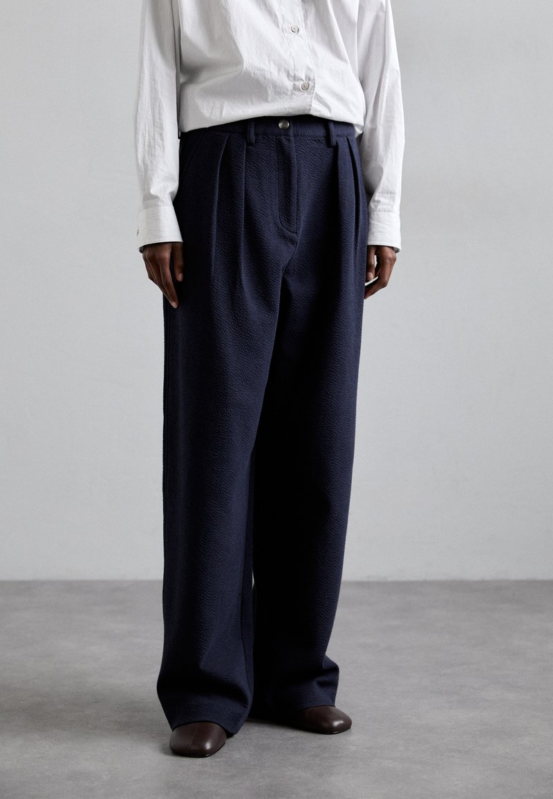 Pantaloni larghi di un blu marina scuro realizzati in tessuto testurizzato con pieghe e chiusura a bottoni, abbinati a una camicia bianca con bottoni e scarpe marroni.