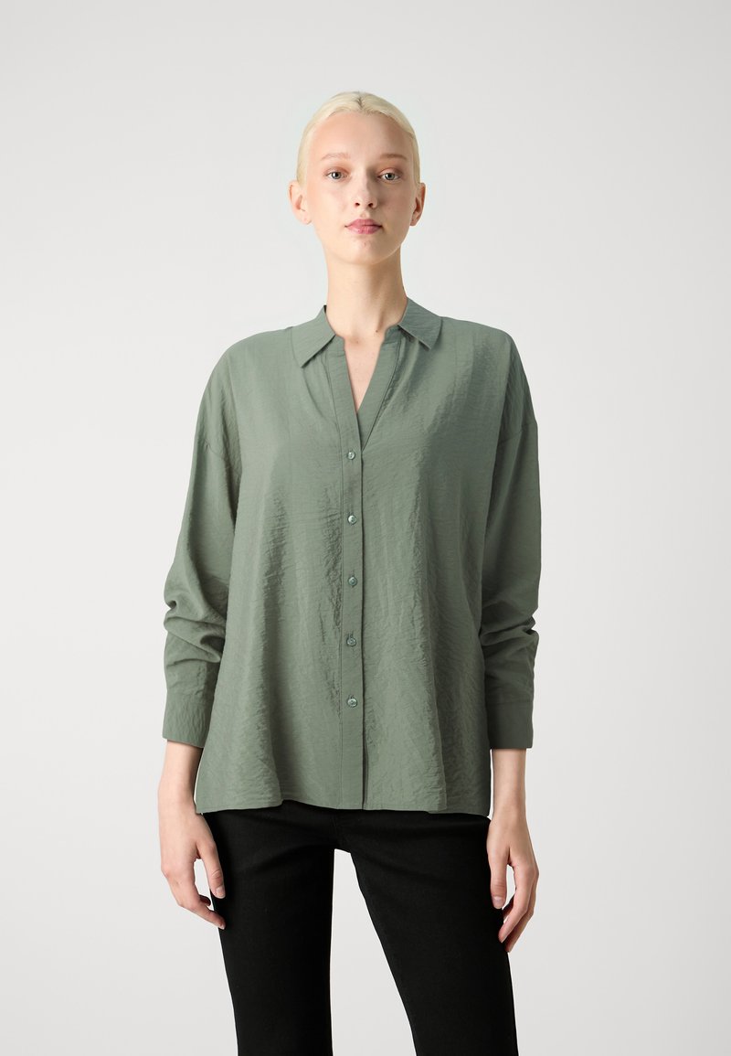 Camisa verde de manga longa com botões, feita de um tecido texturizado e com colarinho. Apresenta um ajuste relaxado e botões contrastantes. Usada com calças pretas.
