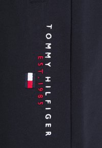 Tissu bleu marine avec texte brodé vertical blanc et rouge « TOMMY HILFIGER EST. 1985 » et petit logo rectangulaire rouge, blanc et bleu.