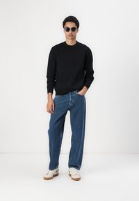 Hanorac negru cu manșete cu nervuri, blugi largi de denim albaștri și adidași albi cu talpă cafenie; persoană purtând ochelari de soare.