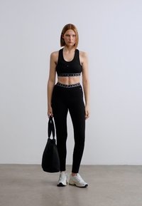 Μαύρο αθλητικό σουτιέν και κολάν με λογότυπο στη ζώνη, συνδυασμένα με λευκά αθλητικά παπούτσια και μαύρη τσάντα tote. Το μοντέλο στέκεται μπροστά σε απλό φόντο.