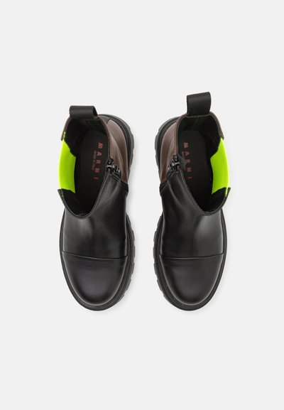 Stivali ankle in pelle nera con un accento in pelle marrone, una vivace fodera interna verde e una suola in gomma testurizzata con chiusura a zip.