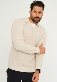 Beige gestrickter Pullover mit Polokragen, gerippten Bündchen und Saum, der ein strukturiertes Muster aufweist. Trägt ein weißes Unterhemd und eine Uhr.