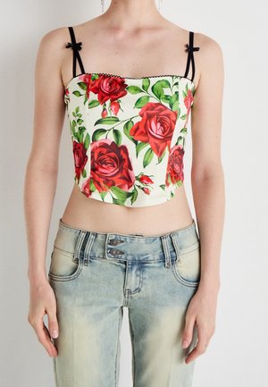 Top corto floral con rosas rojas y hojas verdes sobre un fondo crema. Detalles de lazos negros en las tiras. Combinado con jeans de mezclilla azul claro envejecidos.