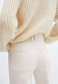 Jean taille haute beige avec deux poches arrière, associés à un pull en tricot crème clair texturé. Design doux et minimaliste.