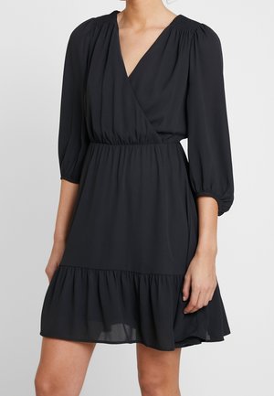 Nainen pukeutuneena mustaan polvipituiseen mekkoon, jossa on 3/4-pituiset pussihihat, v-aukollinen kietaisupääntie, joustava vyötärö ja röyhelöhelma, seisomassa yksiväristä taustaa vasten.