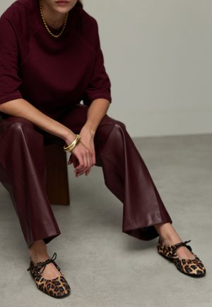 Pantalons larges en cuir bordeaux associés à un haut bordeaux et des ballerines à imprimé léopard avec des accents en satin noir. Un bracelet en or est visible.