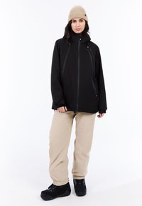 Zwarte waterdichte jas met een hoge kraag, verstelbare capuchon en ritszakken, gecombineerd met beige broek en zwart schoeisel.