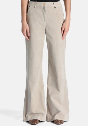 Pantalon beige en velours côtelé à jambes larges avec poches avant et boutons dorés, porté avec des chaussures à motif peau de serpent.