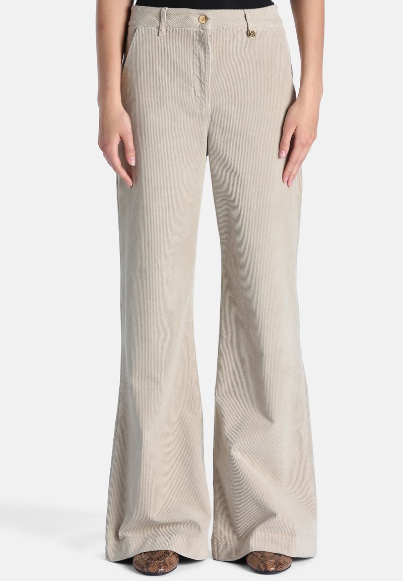 Pantalon beige en velours côtelé à jambes larges avec poches avant et boutons dorés, porté avec des chaussures à motif peau de serpent.