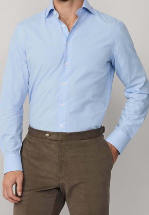 Uomo che indossa una camicia azzurro chiaro a maniche lunghe con bottoni e pantaloni marroni con una mano in tasca, su sfondo semplice.
