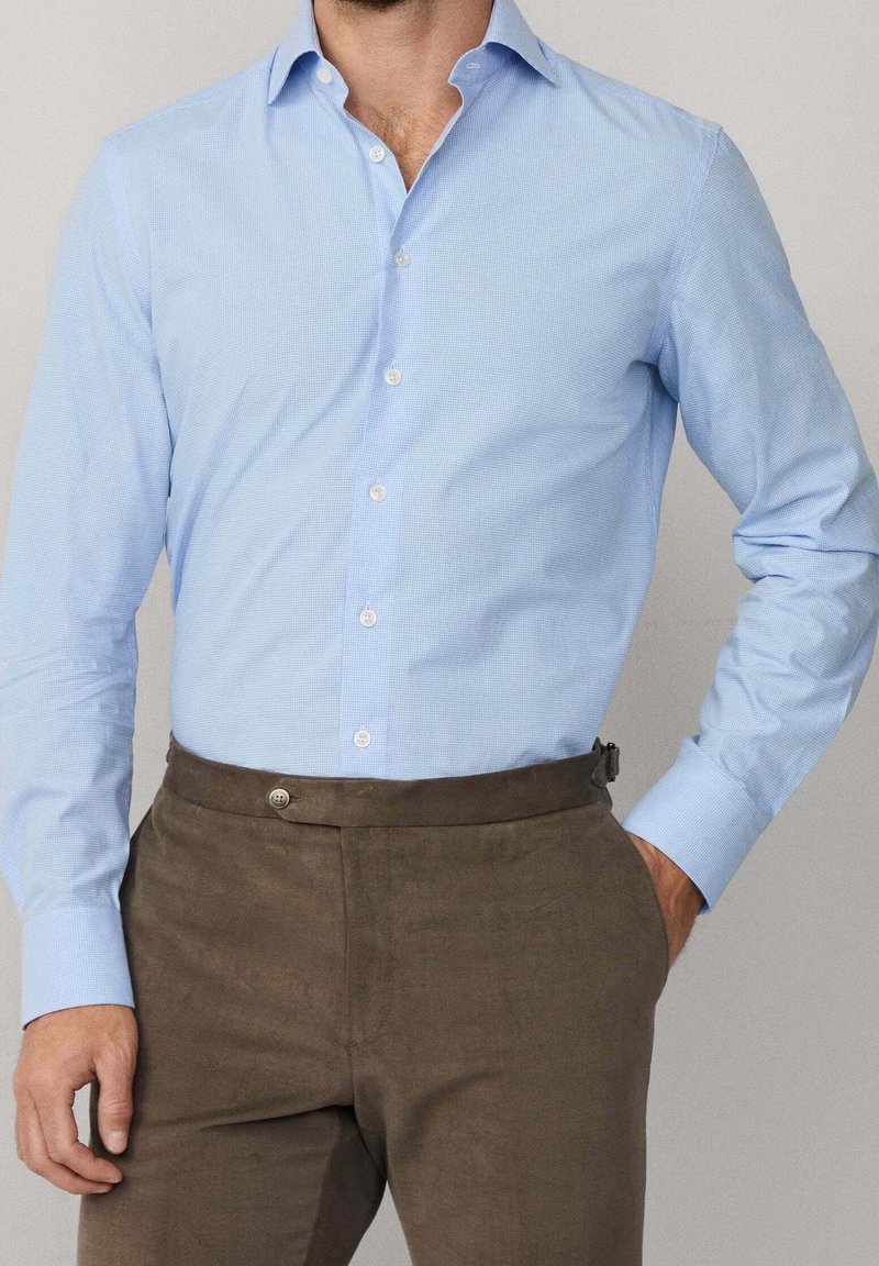 Homme portant une chemise bleue claire à manches longues boutonnée et un pantalon marron, une main dans la poche, sur un fond uni.