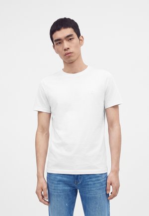 Jeune homme portant un t-shirt blanc uni à manches courtes et un jean bleu debout devant un fond gris clair.