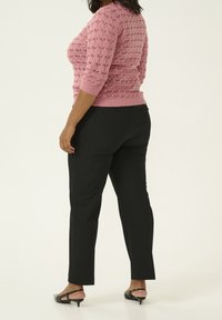 Rosa, strukturiertes Strickoberteil mit Dreiviertelärmeln, kombiniert mit schwarzen Hosen und schwarzen Absatzsandalen. Der neutrale Hintergrund hebt den Fokus auf das Outfit.