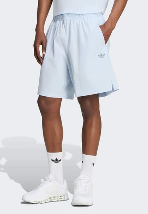 Mand iført lyseblå Adidas-shorts, hvide Adidas-strømper og hvide atletikskor står med den ene hånd i lommen.