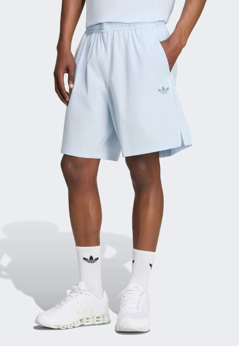 Hombre con pantalones cortos Adidas azul claro, calcetines Adidas blancos y zapatillas deportivas blancas, de pie con una mano en el bolsillo.