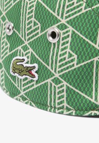 Cappello di tessuto verde con motivo geometrico in crema chiaro, dotato di occhielli in argento e logo di coccodrillo ricamato.