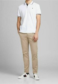 Maglietta polo bianca con strisce nere sul colletto, abbinata a pantaloni beige e sneakers bianche con dettagli rossi e navy. Design semplice.