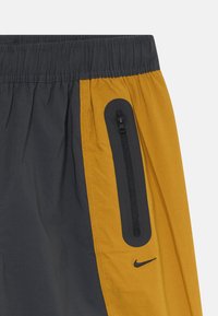 Tuvumais redzams melniem un sinepju dzelteniem sporta šortiem ar elastīgu jostu, vertikālu rāvējslēdzēja kabatu un nelielu Nike swoosh logotipu uz sinepju dzeltenā paneļa.