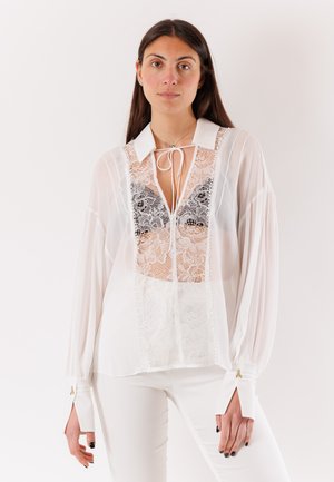 Donna che indossa una camicetta bianca trasparente con pannelli frontali in pizzo sopra un reggiseno nero, abbinata a pantaloni bianchi, su uno sfondo chiaro uniforme.