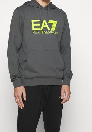 Tummanharmaa huppari neonkeltaisella "EA7 Emporio Armani"-logolla, jota käyttää henkilö mustissa housuissa valkoisella taustalla.
