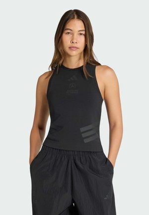 Vrouw draagt een zwarte mouwloze top en losse zwarte broek, staand met handen in de zakken tegen een effen lichte achtergrond.