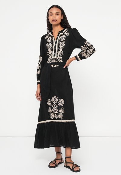 Fekete, hosszú ujjú ruha fehér virágmintás hímzéssel, gombos dekoltázs és bővülő szegéllyel, lapos fekete szandálokkal párosítva.