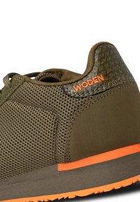 Maslinasto zelena tenisica s mrežastim gornjim dijelom, teksturiranim bočnim panelom, narančastim naglaskom na potplatu i "WODEN" logotipom na stražnjoj strani. Guma potplat s uzorkom za prianjanje.