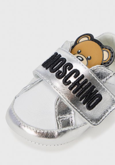 Silvriga metalliska sneakers med en kardborreband med ordet "Moschino" och en björndetalj på tungan, tillverkade av syntetmaterial.