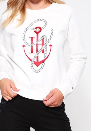 Biała bluza z grafiką czerwonego kotwicy i liny oraz czarne spodnie, pokazane od ramion do bioder, dłonie trzymające rant bluzy.