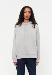 Vrouw met krullend haar, gekleed in een lichtgrijze hoodie en donkerblauwe broek, staande tegen een witte achtergrond en recht vooruit kijkend.