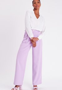 Blouse blanche avec un motif de pois argentés et des poignets volantés, assortie à un pantalon taille haute lavande et des sandales à talons roses pâles.