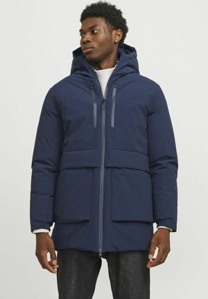 Marineblauer Pufferjacke mit Reißverschluss, großen Vorderentaschen und verstellbarem Kapuzen. Das Material wirkt glatt und isoliert für Wärme.