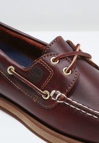 Chaussure bateau en cuir marron foncé avec coutures couleur fauve, œillets métalliques, lacets en cuir et semelle en caoutchouc marron clair sur fond blanc.