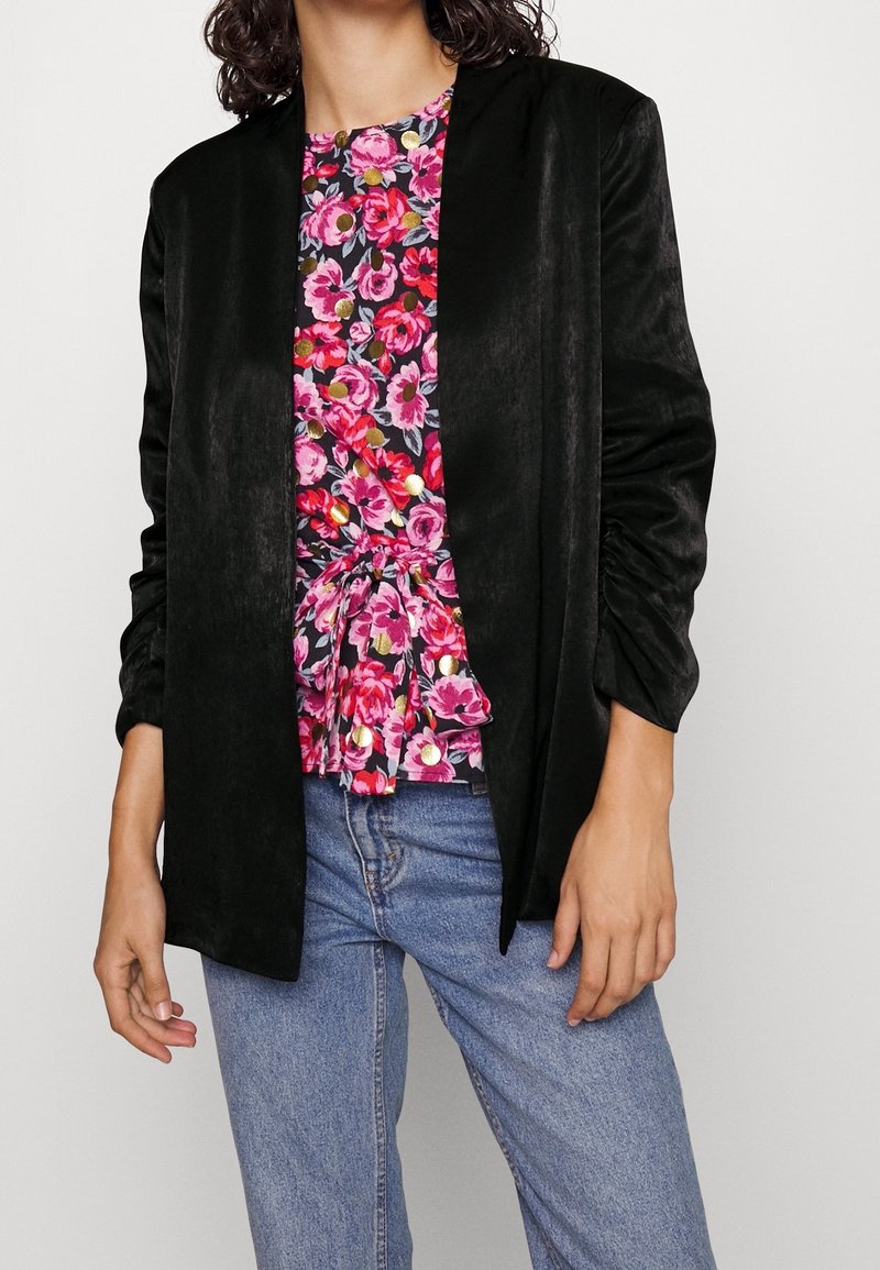 Personne portant une veste noire ouverte sur un chemisier rose fleuri avec une ceinture nouée à la taille et un jean bleu sur un fond uni.