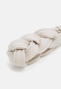 Geflochtener Handtaschenriemen aus weichem, strukturiertem weißen Leder, mit sichtbaren Nähten und einer abgerundeten Form, die das flexible Design betont.