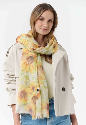 Woman wearing beige trench coat and light yellow floral scarf with sunflower pattern, paired with light yellow top and blue jeans.