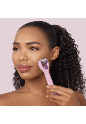 Kvinde med krøllet hår bruger en lyserød dermarulle på sin højre kind mod en ensfarvet lys lilla baggrund.