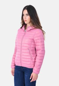Donna con lunghi capelli castani che indossa una giacca trapuntata rosa con zip anteriore e jeans blu, in piedi su uno sfondo bianco.