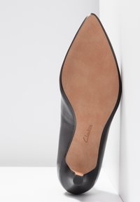 Chaussure en cuir noir à bout pointu vue par la semelle, avec une semelle en cuir lisse couleur beige et le logo "Clarks" près du talon.