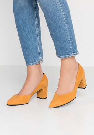 Pieds portant un jean bleu et des chaussures en daim jaune moutarde à talon bloc avec des bouts pointus sur un sol blanc.
