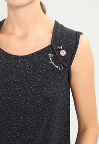 Bardzo ciemnoszary top bez rękawów z małymi białymi i różowymi kropkami, tekst "Reserved" w pobliżu dekoltu, różowy guzik i czarna przypinka na ramieniu.