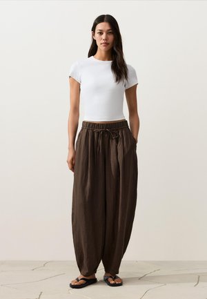 Femme debout portant un haut blanc ajusté à manches courtes, un pantalon marron ample avec cordon de serrage et des sandales noires sur un sol en pierre clair.