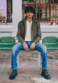 Olīvzaļš žakets virs baltas krekliņa, gaiši zili džinsi, melni šņorējami zābaki ar struktūrizētām zoles, sēž uz pelēka sola starp zaļām krēslām.