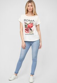 Valkoinen lyhythihainen t-paita, jossa on granaattiomenia kuvaava grafiikka ja sana "ROMA" mustalla, yhdistettynä vaaleansinisiin slim fit -farkkuihin ja valkoisiin lenkkareihin.