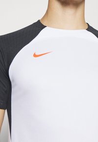 Camiseta deportiva blanca con mangas gris oscuro, que presenta un diseño texturizado. Logo de Nike naranja destacado en el pecho.
