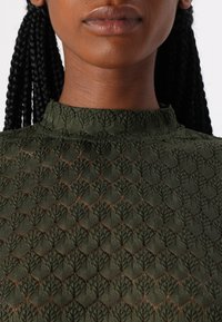 Žalia siuvinėta palaidinė su aukštu kaklu, kuri turi tekstūrinį raštą su ventiliuojančiomis formomis visame audinyje.