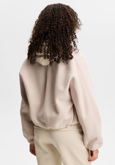 Person with curly hair wearing a light beige fleece hoodie and matching pants, shown from the back against a plain background.