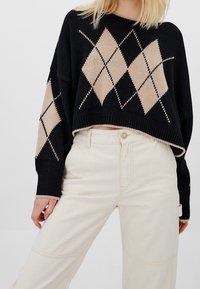Suéter corto negro con patrón de rombos beiges y puños acanalados; combinado con pantalones de pierna ancha color crema. Textura de tejido de punto suave.