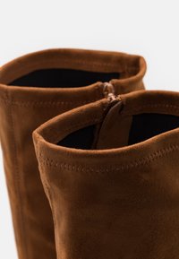 Bruna mockakängor som går upp till knäna med en slät yta, med en dragkedja högst upp och en sömnadsdetalj längs kragen.